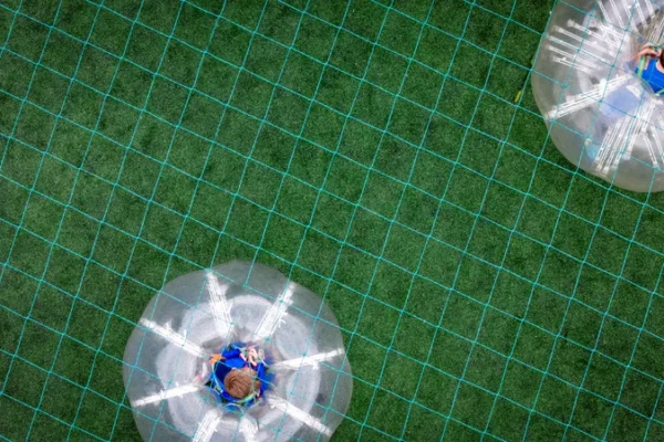 Overhead view of two people playing bubble soccer on a green field with a grid pattern. Each person is inside an inflated, transparent ball.